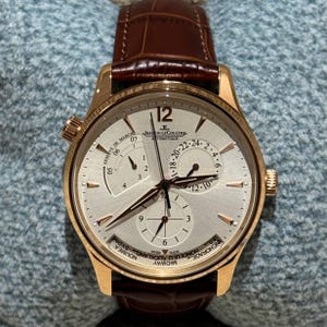 Op de afbeelding: Een goudkleurig Jaeger-LeCoultre automatisch polshorloge met een bruine leren band. De zilveren wijzerplaat heeft meerdere sub-wijzerplaten en wijzers, die de tijd, datum en gangreserve weergeven. De wijzerplaat heeft de tekst "Jaeger-LeCoultre" en "AUTOMATIQUE".