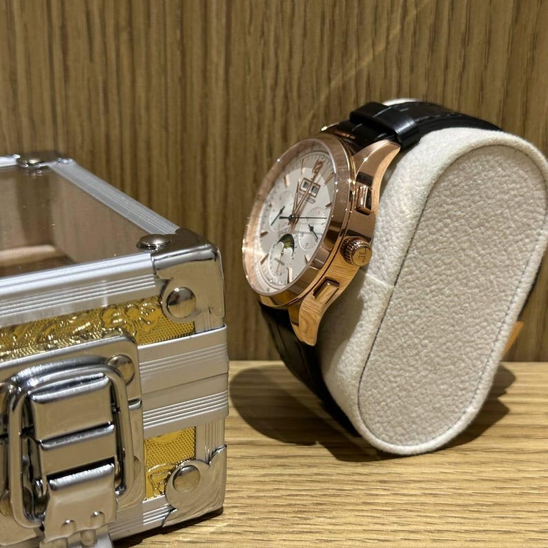 May include: A rose gold wristwatch with a white face and black leather band is displayed on a cream-colored watch stand. The watch has a complex dial with multiple sub-dials. A silver and gold jewelry box is partially visible in the foreground.