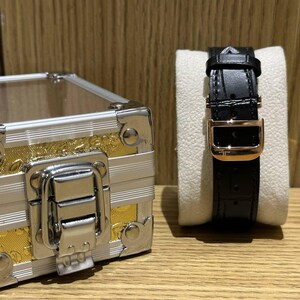 May include: A black leather watch band with a gold-colored buckle is displayed on a white watch stand. A small silver and gold-colored decorative box is in the background.
