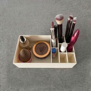 Könnte beinhalten: Ein cremefarbener Make-up-Organizer mit verschiedenen Schönheitsprodukten. Enthält Make-up-Pinsel, einen braunen Puder-Kompakt mit dem Text "CITY BRONZER", einen braunen Make-up-Schwamm und Make-up-Fläschchen.