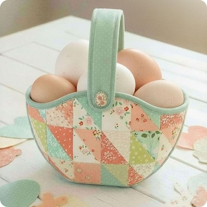 Op de afbeelding: Een patchworkmand in pastelkleuren gevuld met eieren. De mand heeft een mintgroene handgreep en is versierd met bloemen- en geometrische patronen in roze, perzik en groen. De eieren zijn een mix van wit en lichtbruin.