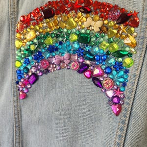May include: A light blue denim jacket featuring a large, arched rainbow design on the back. The rainbow is constructed from an array of multicoloured, faceted plastic gems, rhinestones, and small decorative charms in various shapes and sizes.
