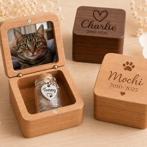 Puede incluir: Cajas conmemorativas de madera para mascotas. Una caja tiene una foto de un gato y un pequeño frasco de vidrio con una etiqueta en forma de corazón que dice "Tommy". Otras dos cajas están cerradas, una marrón oscuro con "Charlie" y la otra marrón claro con "Mochi".