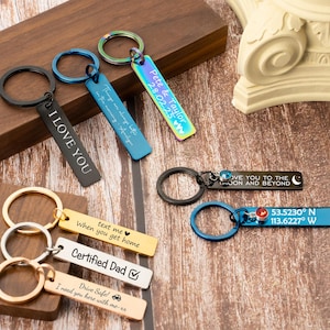 Peut inclure: Plusieurs porte-clés personnalisés de différentes couleurs, dont noir, bleu, or et irisé. Certains porte-clés comportent des textes tels que "I LOVE YOU", "Certified Dad" et des coordonnées. Les porte-clés sont sur une surface en bois.