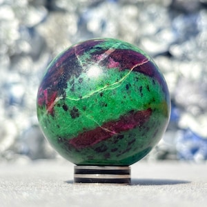 Könnte beinhalten: Eine polierte Kugel aus Rubin-Zoisit, die eine lebendige Mischung aus Grün, tiefem Rot und Schwarz aufweist. Der Edelsteinball ruht auf einem silber-schwarzen Sockel und zeigt seine natürlichen Muster. Ein einzigartiger Dekorationsgegenstand.