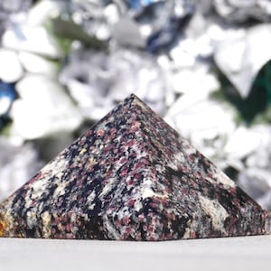 May include: A pyramid-shaped decorative object with a speckled pattern of dark red, black, and white. The pyramid has a flat base and a pointed top. The background is blurred with white and blue tones.