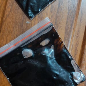 Könnte beinhalten: Zwei transparente Plastiktüten mit rotem Reißverschluss, die jeweils eine dunkle, zähflüssige Substanz enthalten. Die Tüten liegen auf einer Holzoberfläche mit warmem, braunem Ton.
