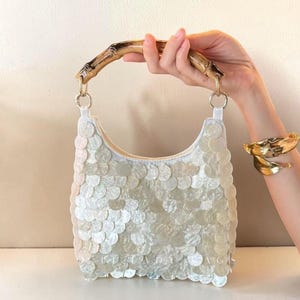 May include: A white handbag covered in overlapping circular iridescent shell-like discs, featuring a curved bamboo handle attached with gold-toned metal rings. The bag has a structured, semi-circular silhouette with a clean, minimalist aesthetic.
