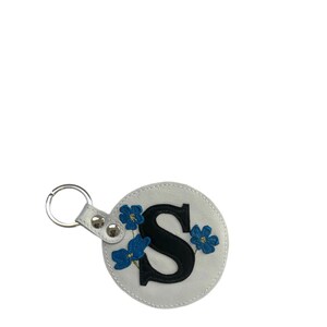 Könnte beinhalten: Weißer Leder-Schlüsselanhänger mit schwarzem Buchstaben "S" und blauer Blumenstickerei. Der runde Schlüsselanhänger hat einen silbernen Schlüsselring und einen weißen Lederriemen mit silbernen Druckknöpfen.