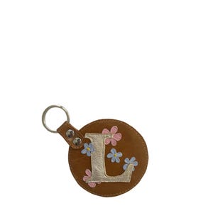Könnte beinhalten: Ein brauner Leder-Schlüsselanhänger mit einem silbernen Metallring. Der Schlüsselanhänger zeigt den Buchstaben "L" in Silber, umgeben von rosa und blauen Blumenapplikationen. Der runde Schlüsselanhänger hat einen Lederriemen mit Metallnieten.