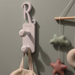 Peut inclure: Un crochet mural blanc avec la lettre "R" et le nom "Raphaël" est fixé sur un mur vert clair. Un sac fourre-tout beige est suspendu au crochet. Un mobile avec des formes en feutre, dont un nuage, une étoile et un anneau, est à droite.