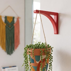 Puede incluir: Una maceta colgante de terracota con un diseño geométrico colorido, suspendida de un soporte de madera rojo. La maceta está llena de una planta suculenta colgante. Un tapiz de macramé está en el fondo.