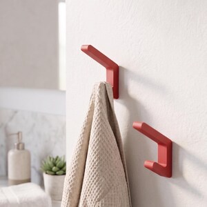 Op de afbeelding: Twee matte rode wandhaken met een hoekig, geometrisch ontwerp. Aan één haak hangt een beige handdoek met wafelstructuur. De haken zijn bevestigd aan een witte muur. De tekst "Lignes / Rouges" is zichtbaar in de rechterbenedenhoek.
