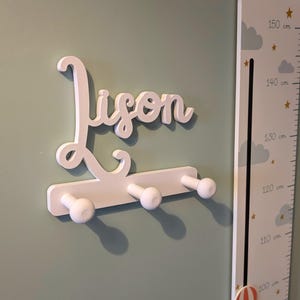Può includere: Un appendiabiti da parete bianco con il nome Lison scritto in corsivo. L'appendiabiti presenta una base orizzontale con tre ganci arrotondati che sporgono in avanti. L'oggetto è fissato su una parete verde chiaro accanto a un metro a muro.