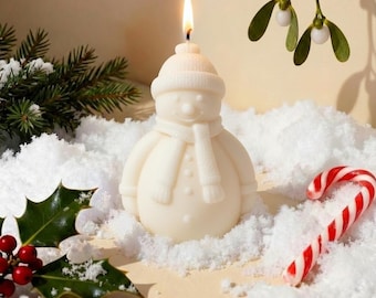 May include: A snowman-shaped candle with a lit wick, wearing a hat and scarf. The candle is white and sits in artificial snow, with a candy cane and sprigs of greenery. Festive holiday decor.