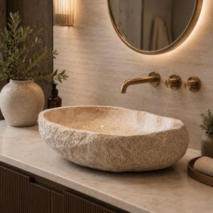 May include: An oval-shaped vessel sink carved from natural beige stone with a rough, textured exterior and a smooth interior basin. The sink is mounted on a light-colored countertop beneath a wall-mounted brass faucet and two matching circular knobs.
