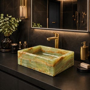 May include: A rectangular vessel sink crafted from polished green onyx stone with natural brown veining. The sink is positioned on a dark countertop beneath a gold-toned faucet, complemented by a matching gold soap dispenser and a black candle.