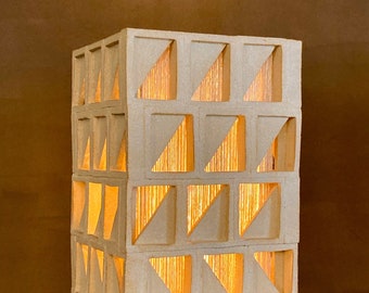 May include: A white ceramic lamp with a geometric design. The lamp has a square base and a rectangular body with a grid of squares and triangles. The light shines through the openings in the design, creating a warm glow.
