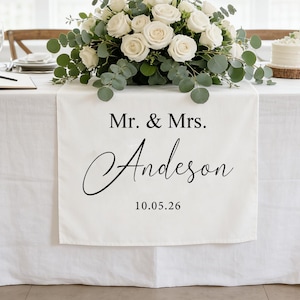 May include: A wedding reception table set with a white tablecloth, floral arrangement, cake, and candles. A banner displays "Mr. & Mrs. Anderson" and the date "10.05.26". The table is set near a large window with natural light.