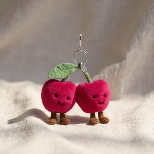 Op de afbeelding: Een pluche kersen sleutelhanger met twee rode kersen met lachende gezichten, bruine voetjes en een groen blad. De sleutelhanger heeft een zilveren ring om aan sleutels of tassen te bevestigen. De kersen zijn zacht en knuffelig.