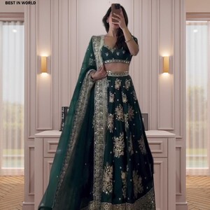 Könnte beinhalten: Ein dunkelgrünes Abendkleid mit Gold- und Silberverzierungen. Das Kleid hat ein Crop-Top, einen langen Rock und einen passenden, durchsichtigen Schal. Das Outfit ist mit aufwendigen Blumenmustern und metallischen Verzierungen versehen. Der Text "BEST IN WORLD" ist sichtbar.