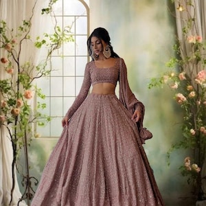 Puede incluir: Un vestido formal de color rosa polvoriento con intrincados bordados. El conjunto incluye un top corto de manga larga y una falda fluida. El atuendo se complementa con grandes pendientes dorados. El fondo presenta una ventana y arreglos florales.