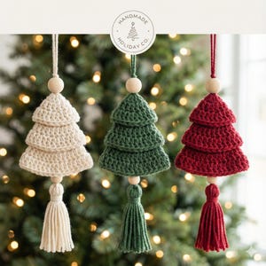May include: Three handmade crochet Christmas tree decorations in cream, green, and red. Each tree has three tiers, a wooden bead, and a matching tassel. The ornaments are suspended in front of a blurred Christmas tree background. The top of the image has a circular logo that says "Handmade Holiday Co."