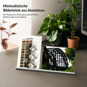 Pode incluir: Uma barra de fotos de alumínio prateado segurando duas pequenas impressões. As impressões apresentam um edifício com uma escada em espiral e uma ponte preta. O texto na imagem diz "Minimalistische Bilderleiste aus Aluminium."