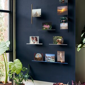 Op de afbeelding: Een display van ingelijste foto's gemonteerd op een donkerblauwe muur. De foto's zijn gerangschikt op kleine planken en verticale metalen staven. De afbeeldingen tonen verschillende scènes, waaronder landschappen, architectuur en mensen. Potplanten staan op de voorgrond.
