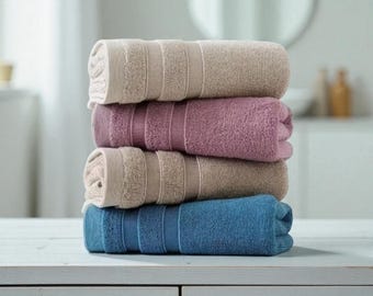 Può includere: Una pila di quattro asciugamani da bagno arrotolati nei toni del beige, malva e blu. Gli asciugamani sono ordinatamente disposti, con strisce strutturate visibili. L'ambientazione è in un bagno, suggerendo tessuti per la casa.