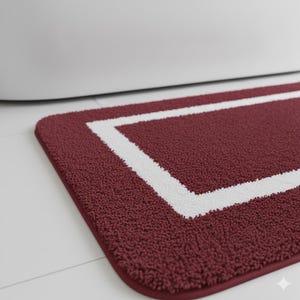 Può includere: Un tappetino da bagno bordeaux con un bordo rettangolare bianco. Il tappetino ha una superficie morbida e strutturata ed è posizionato su un pavimento bianco vicino a una vasca da bagno bianca.