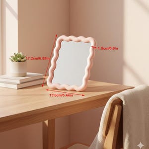 Peut inclure: Un miroir rose aux bords ondulés, de 17,2 cm de haut et 13,6 cm de large, posé sur une surface en bois. Le cadre du miroir est rose clair. Une petite plante succulente dans un pot blanc et une pile de livres sont visibles en arrière-plan.