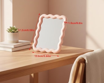 May include: A pink, wavy-edged mirror, 6.88 inches tall and 5.44 inches wide, stands on a wooden surface. The mirror's frame is a soft pink color. A small succulent in a white pot and a stack of books are visible in the background.