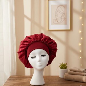 Peut inclure: Un bonnet en satin bordeaux, présenté sur une tête de mannequin blanche. Le bonnet a une large bande et une couronne froncée. Le mannequin est sur une table en bois, avec une petite plante et des serviettes en arrière-plan. L'éclairage est doux et chaleureux.