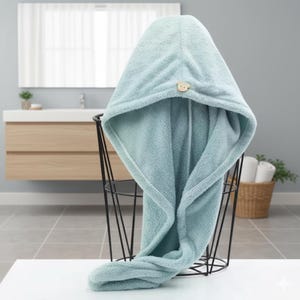 Könnte beinhalten: Hellblaues Kapuzen-Haarhandtuch mit einem hölzernen Bärenknopf. Das Handtuch besteht aus einem weichen, saugfähigen Material und ist über einem schwarzen Drahtkorb drapiert. Es ist zum Einwickeln der Haare nach dem Waschen gedacht.