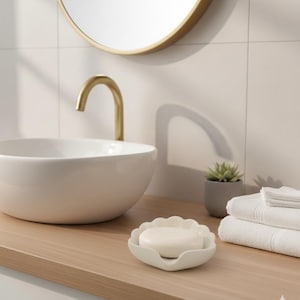 以下が含まれることがあります： 木製のカウンタートップに、白いスカラップエッジの石鹸皿が石鹸を保持しています。白いセラミックシンク、金の蛇口、丸い鏡が背景にあります。小さな鉢植えの植物と折りたたまれた白いタオルも表示されています。