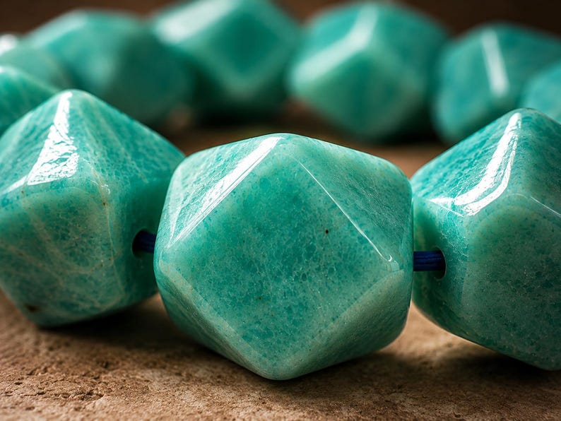 Pu&ograve; includere: Una collana di perline di amazzonite verde acqua sfaccettate e lucidate, infilate su un sottile cordino blu. Ogni perla presenta una forma geometrica a pi&ugrave; facce con una superficie liscia e lucida che riflette la luce, mostrando le sfumature.