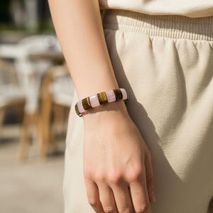 May include: A close-up of a bracelet featuring alternating rectangular beads of pink, brown, and tan. The bracelet is worn on a person's wrist, with the hand and arm visible. The background is blurred, suggesting an outdoor setting.