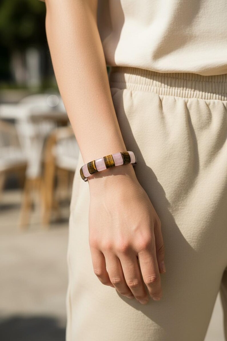 May include: A close-up of a bracelet featuring alternating rectangular beads of pink, brown, and tan. The bracelet is worn on a person's wrist, with the hand and arm visible. The background is blurred, suggesting an outdoor setting.