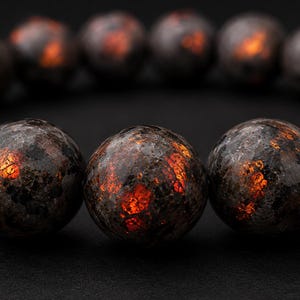 Può includere: Un bracciale composto da perline sferiche con una superficie scura e strutturata nei toni del marrone scuro e del nero. Ogni perlina presenta riflessi interni di luce arancione e rossa vibrante che sembrano brillare sulla finitura in pietra.