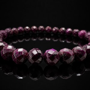 Op de afbeelding: Een close-up van een robijn armband tegen een zwarte achtergrond. De armband heeft talrijke gefacetteerde, dieprode edelstenen. De edelstenen zijn rond en variëren in grootte, waardoor een textuur ontstaat. Het licht reflecteert op de facetten.