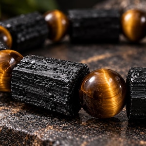 Könnte beinhalten: Ein Armband mit abwechselnd schwarzen, rau texturierten, zylindrischen Turmalinperlen und glatten, polierten, goldbraunen Tigeraugen-Kugeln. Das Design zeigt einen Kontrast zwischen dem rohen, mattschwarzen Stein und den reflektierenden, schimmernden Oberflächen der braunen Edelsteine.