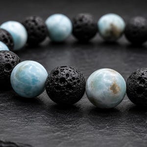 Puede incluir: Una pulsera de cuentas con esferas alternas de piedra lisa de color azul claro veteado y roca volcánica negra mate porosa. Las cuentas están dispuestas en un patrón repetitivo, creando un contraste entre las superficies azules pulidas y las esferas negras texturizadas.