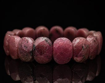 May include: A close-up of a ruby bracelet. The bracelet is made of oval-shaped, polished ruby stones. The stones are a deep pinkish-red color with some darker inclusions. The bracelet is set against a black background.