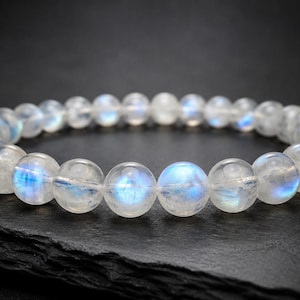 Op de afbeelding: Een cirkelvormige armband bestaande uit gladde, ronde, doorschijnende maansteen kralen. Elke kraal vertoont een uitgesproken iriserende blauwe adularescentie, die het licht reflecteert met een zachte gloed. De kralen zijn in een lus geregen.
