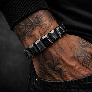 Op de afbeelding: Een zwarte armband met een geometrisch ontwerp. De armband is gemaakt van rechthoekige, facetgeslepen schakels. De afbeelding toont de armband om een pols, met tatoeages zichtbaar op de arm en hand. De armband is een opvallend accessoire.