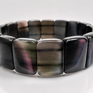 Könnte beinhalten: Ein Stretch-Armband mit rechteckigen, irisierenden Perlen in Schwarz, Grau und Lila. Die polierten Steine haben eine glänzende Oberfläche und sind zu einer durchgehenden Schlaufe aufgereiht. Das Armband wird auf einer weißen Oberfläche präsentiert.