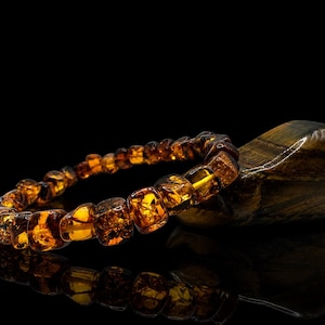 May include: A bracelet made of amber-colored, square-shaped beads. The bracelet is resting on a dark, polished stone. The amber beads have a warm, golden hue, and the stone has dark brown and gold stripes. The background is black.