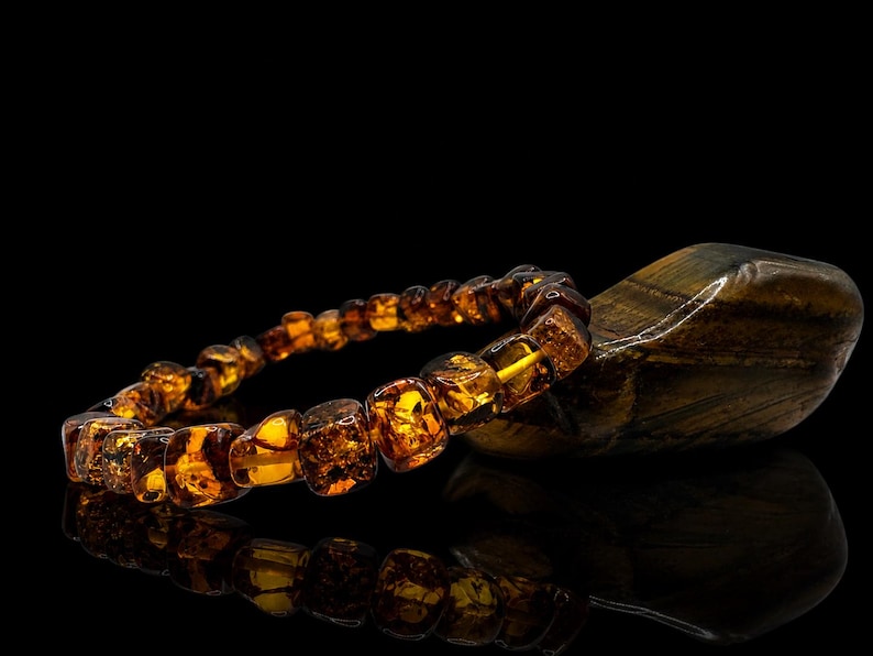 May include: A bracelet made of amber-colored, square-shaped beads. The bracelet is resting on a dark, polished stone. The amber beads have a warm, golden hue, and the stone has dark brown and gold stripes. The background is black.