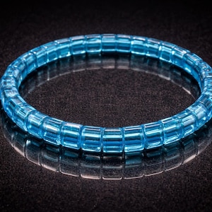 Op de afbeelding: Een lichtblauwe armband bestaande uit rechthoekige, transparante kralen. De armband is cirkelvormig en reflecteert op een donkere, getextureerde ondergrond. De kralen zijn in een doorlopende lus gerangschikt, wat een strak ontwerp creëert. De kleur van de armband is helder blauw.